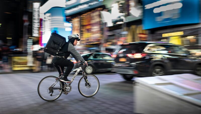 大きな配達バッグを背負い都市部を自転車で走るフードデリバリー配達員
