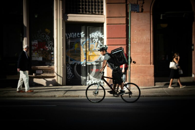 大型の配達バッグを背負い市街地を自転車で走るフードデリバリー配達員