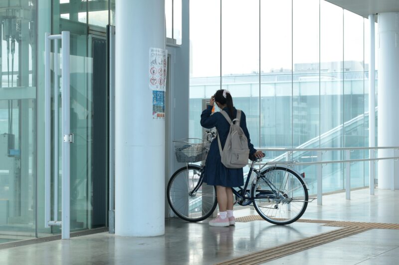 リュックを背負った女性が建物内で前かご付き自転車を押している様子