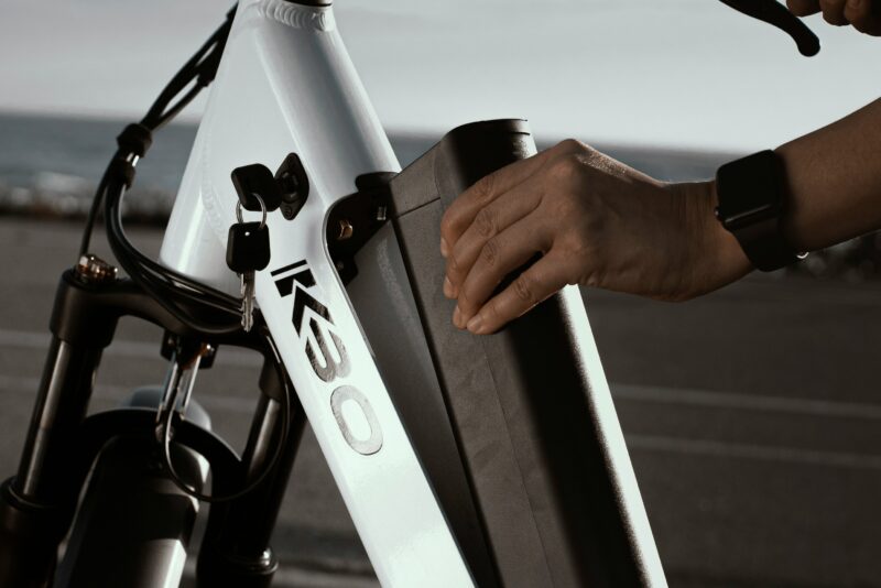 白いKBOの電動アシスト自転車からバッテリーを取り外している様子。フレームのバッテリー装着部分と鍵穴が見え、充電前の取り外し方法を説明する写真。